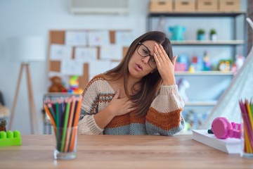 Canvas Print - Young beautiful teacher woman wearing sweater and glasses sitting on desk at kindergarten Touching forehead for illness and fever, flu and cold, virus sick