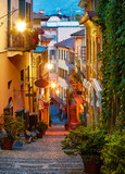 Fototapeta Uliczki - Bellagio village at lake Como near Milan Italy, region Lombardy. Famous street with paving stones stairs and cosy restaurants during sunrise with glowing lanterns and green plants on old houses walls.