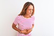 Young redhead woman wearing striped casual t-shirt stading over white isolated background with hand on stomach because indigestion, painful illness feeling unwell. Ache concept.