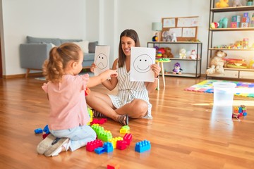 Wall Mural - Beautiful psycologist and blond toddler girl sitting on the floor doing therapy using emoji emotions at consulting room