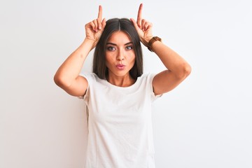 Young beautiful woman wearing casual t-shirt standing over isolated white background doing funny gesture with finger over head as bull horns