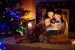 © MNStudio - Happy young mother and her two small daughters opening a magical Christmas gift by a fireplace in a cozy dark living room on Christmas eve.