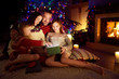 © MNStudio - Happy young family of four using a tablet pc at home by a fireplace in warm and cozy living room on Christmas eve.