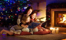 Happy Young Mother And Her Daughters Reading A Story Book Together By A Fireplace In A Cozy Dark Living Room On Christmas Eve. Celebrating Xmas At Home.
