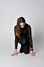 Full Length Portrait Of A Pretty Brunette Woman Wearing Black Leather Fantasy Costume. Crouching Pose On A Studio Background.