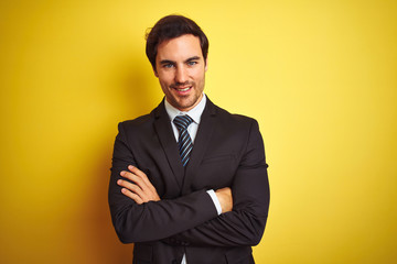 Young handsome businessman wearing suit and tie standing over isolated yellow background happy face smiling with crossed arms looking at the camera. Positive person.