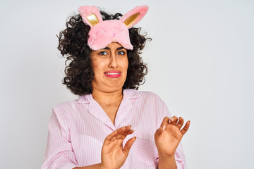 Sticker - Arab woman with curly hair wearing pajama and sleep mask over isolated white background disgusted expression, displeased and fearful doing disgust face because aversion reaction. With hands raised