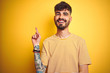 © Krakenimages.com - Young man with tattoo wearing striped t-shirt standing over isolated yellow background with a big smile on face, pointing with hand finger to the side looking at the camera.