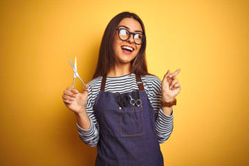 Wall Mural - Young beautiful hairdresser woman holding scissors standing over isolated yellow background very happy pointing with hand and finger to the side