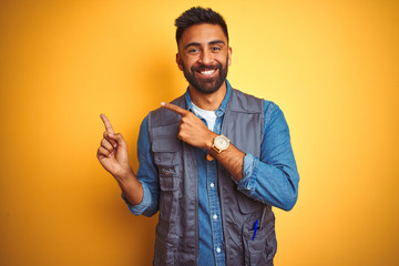 Handsome indian technician man wearing uniform over isolated yellow background smiling and looking at the camera pointing with two hands and fingers to the side.