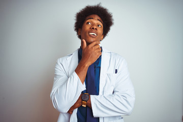 Poster - Young african american doctor man wearing coat standing over isolated white background with hand on chin thinking about question, pensive expression. Smiling with thoughtful face. Doubt concept.