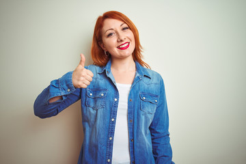 Sticker - Young beautiful redhead woman wearing denim shirt standing over white isolated background doing happy thumbs up gesture with hand. Approving expression looking at the camera with showing success.