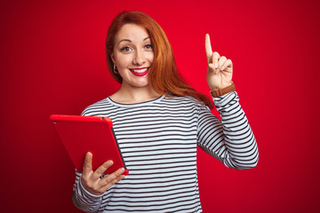 Young beautiful redhead woman using tablet over red isolated background surprised with an idea or question pointing finger with happy face, number one