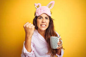 Wall Mural - Young woman wearing pajama and mask drinking coffee over yellow isolated background annoyed and frustrated shouting with anger, crazy and yelling with raised hand, anger concept