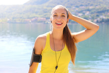 Happy Fitness Girl Runner Listening To Running Motivation Music On Smartphone App With Earphones. Young Woman Athlete Wearing Tank Top And Armband Phone Looking At Camera Outdoor On City Lake.