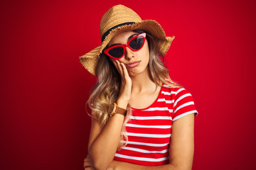 Poster - Young beautiful woman wearing sunglasses and summer hat over red isolated background thinking looking tired and bored with depression problems with crossed arms.