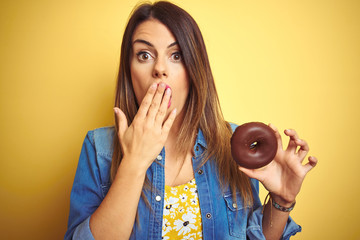 Poster - Young beautiful woman eating chocolate donut over yellow background cover mouth with hand shocked with shame for mistake, expression of fear, scared in silence, secret concept