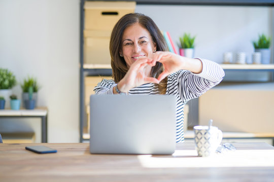 Middle age senior woman sitting at the table at home working using computer laptop smiling in love doing heart symbol shape with hands. Romantic concept.