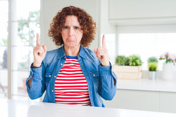 Wall Mural - Middle age senior woman with curly hair wearing denim jacket at home Pointing up looking sad and upset, indicating direction with fingers, unhappy and depressed.