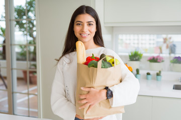 Sticker - Beautiful young woman smiling holding a paper bag full of groceries at the kitchen