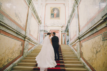 A beautiful young Turkish man wearing a black suit and a beard standing in the home along with his beautiful bride in a white dress and a bouquet of flowers