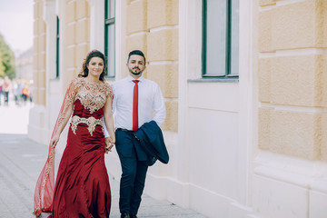 Beautiful woman in a long red dress walks around the city with her husband in a blue suit and with a beard