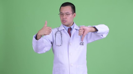 Wall Mural - Confused Asian man doctor with eyeglasses choosing between thumbs up and thumbs down