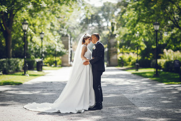 Wall Mural - Elegant bride in a white dress and veil. Handsome groom in a blue suit. Couple in a summer park