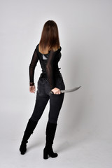 Sticker - full length portrait of a pretty brunette woman wearing black leather fantasy costume. Standing pose, holding a dagger on a studio background.