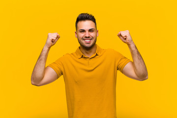 Wall Mural - young hispanic man feeling happy, surprised and proud, shouting and celebrating success with a big smile against orange wall