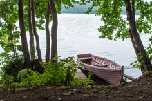Old Abandoned Rotten Unnecessary Small Boat With A Broken Bottom On A Rusty Chain With Oars On The Lake On The Shore With Water Inside, At The Platform Near The Downed Old