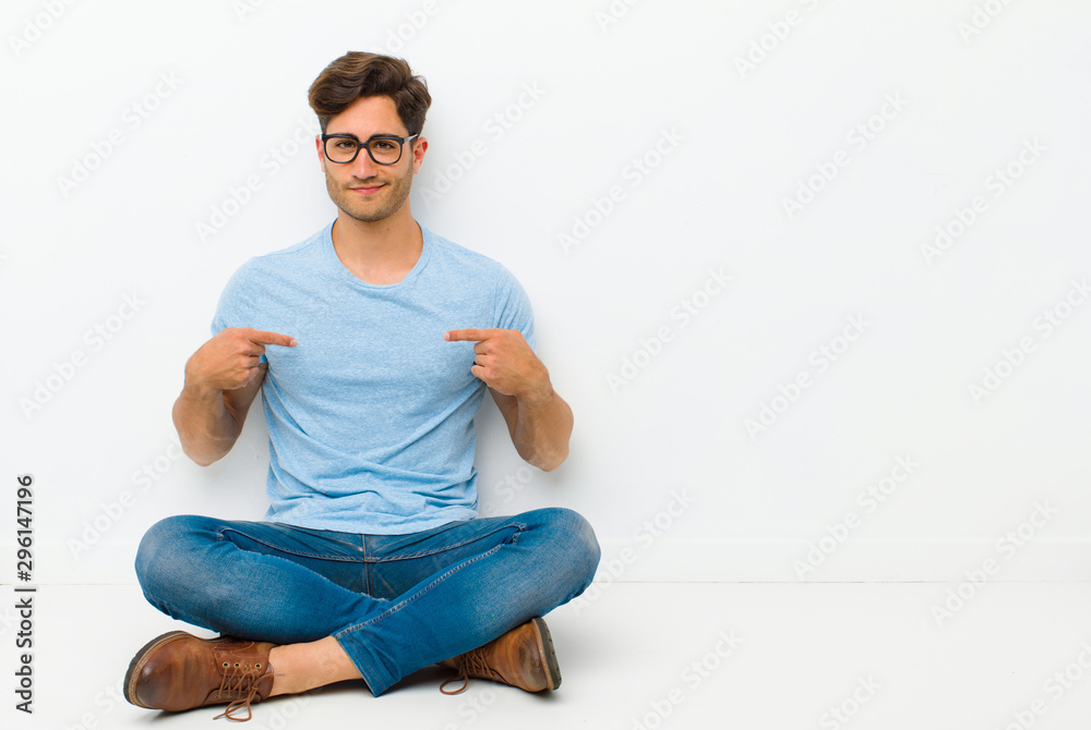 young handsome man looking proud, positive and casual pointing to chest with both hands sitting on the floor - obrazy, fototapety, plakaty 