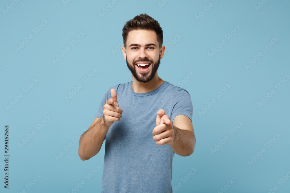 Young cheerful handsome man in casual clothes posing isolated on blue wall background, studio portrait. People sincere emotions lifestyle concept. Mock up copy space. Pointing index fingers on camera. - obrazy, fototapety, plakaty 
