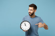 Young handsome man in casual clothes posing isolated on blue background, studio portrait. People lifestyle concept. Mock up copy space. Holding in hands, pointing index finger on clock, looking aside.