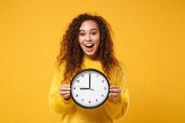 Excited young african american girl in fur sweater posing isolated on yellow orange background, studio portrait. People lifestyle concept. Mock up copy space. Holding clock, keeping mouth wide open.