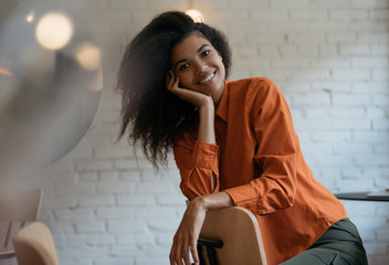 Authentic portrait of young cheerful African American woman with beautiful smiling face, curly hairstyle sitting on chair in modern cafe. Cute fashion model posing for pictures, laughing 