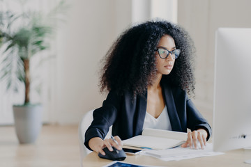 Serious curly businesswoman focused at display of computer, works on making project, surrounded with textbook and papers, wears glasses for vision correction, black suit, poses in office room