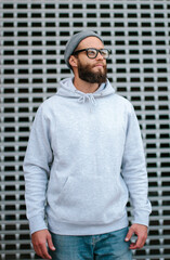 Wall Mural - City portrait of handsome hipster guy with beard wearing gray blank hoodie or sweatshirt and hat with space for your logo or design. Mockup for print