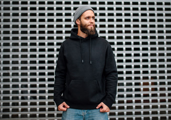 Sticker - City portrait of handsome hipster guy with beard wearing black blank hoodie or sweatshirt and hat with space for your logo or design. Mockup for print