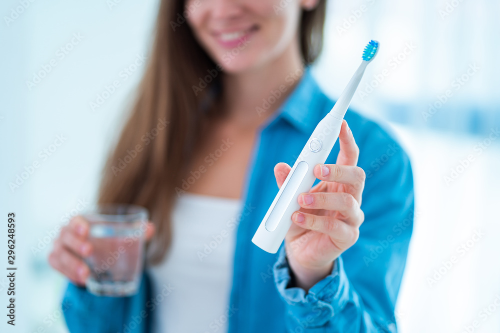 Happy smiling woman with ultrasonic electric toothbrush in bathroom at home. Dental hygiene and oral care, healthy teeth - obrazy, fototapety, plakaty 