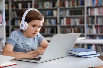 Wall Mural - Little boy with headphones reading book using laptop in library