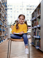 Cute little girl with headphones reading book on chair in library