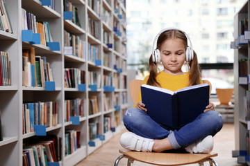 Wall Mural - Cute little girl with headphones reading book on chair in library