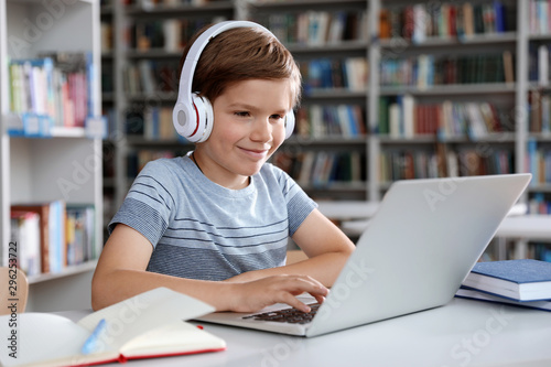 Fotografía Little boy with headphones reading book using laptop in library