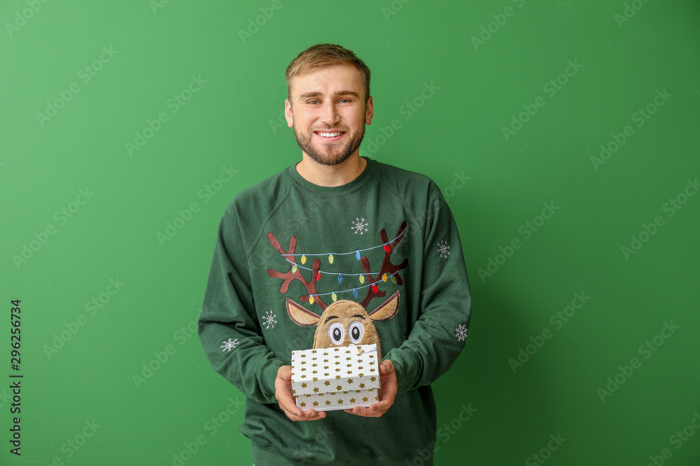 Young man in Christmas sweater and with gift on color background