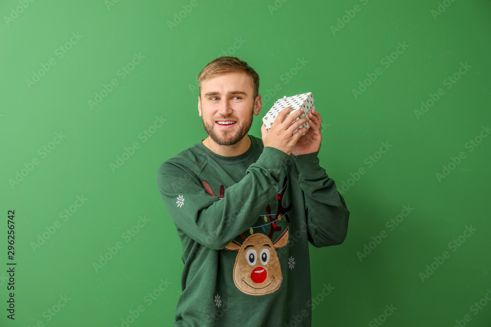 Young man in Christmas sweater and with gift on color background