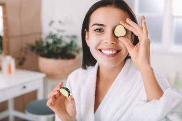 Canvas Print - Beautiful young woman with facial mask and cucumber slices at home