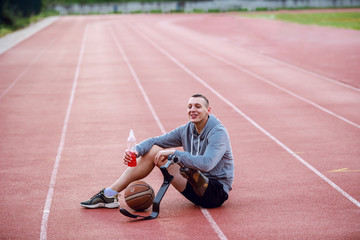 Poster - Highly motivated handsome caucasian sporty handicapped man in sportswear sitting on racetrack and holding refreshment. Between legs is basketball ball.