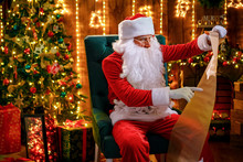 Portrait Of Happy Santa Claus Sitting At His Room At Home Near Christmas Tree, Fireplace And Reading Christmas Letter Or Wish List