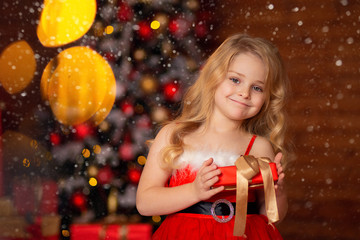 Wall Mural - Beautiful blonde girl in a bright Red festive dress holds a gift in her hands in anticipation of the New Year against the background of a wooden house.